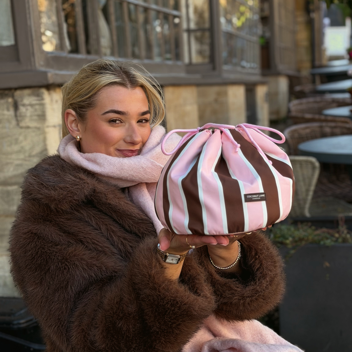 Woman wearing a baby pink scarf and brown faux fur jacket, holding a drawstring toiletry bag featuring chocolate brown, baby pink and white stripes with pink drawstring closure.