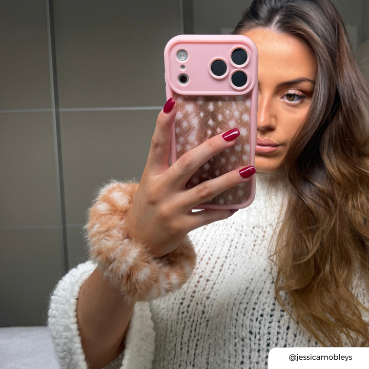 girl in mirror wearing white holding a pink and fawn print phone case, she has a matching fluffy scrunchie on her wrist