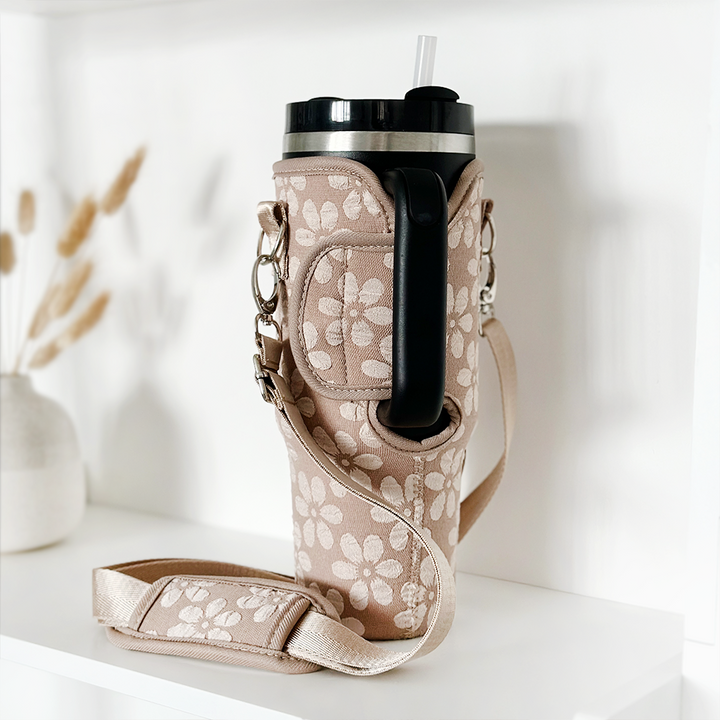The Tumbler Carry Case - Mocha Flower by Coconut Lane cradles a large insulated tumbler with a black lid and straw, its beige fabric adorned with white floral patterns. The holder, complete with a shoulder strap, sits on a white shelf alongside a minimalist vase and dried flowers in the blurred background.