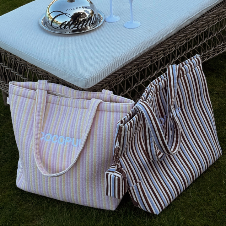 Two pastel striped Cocopup tote bags sit on grass next to a wicker bench with drinks and a serving tray.