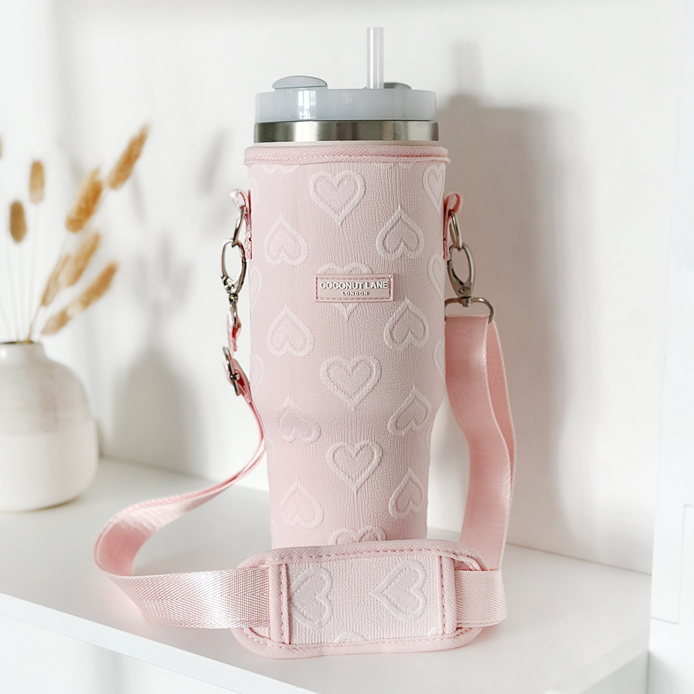 A Tumbler Carry Case - Baby Pink Heart by Coconut Lane sits elegantly on a white surface, featuring an insulated pink design with a heart pattern and an attached shoulder strap for easy carrying. For added convenience, this travel mug case offers excellent tumbler protection. In the background on the left, a vase with dried wheat stalks adds a natural touch.