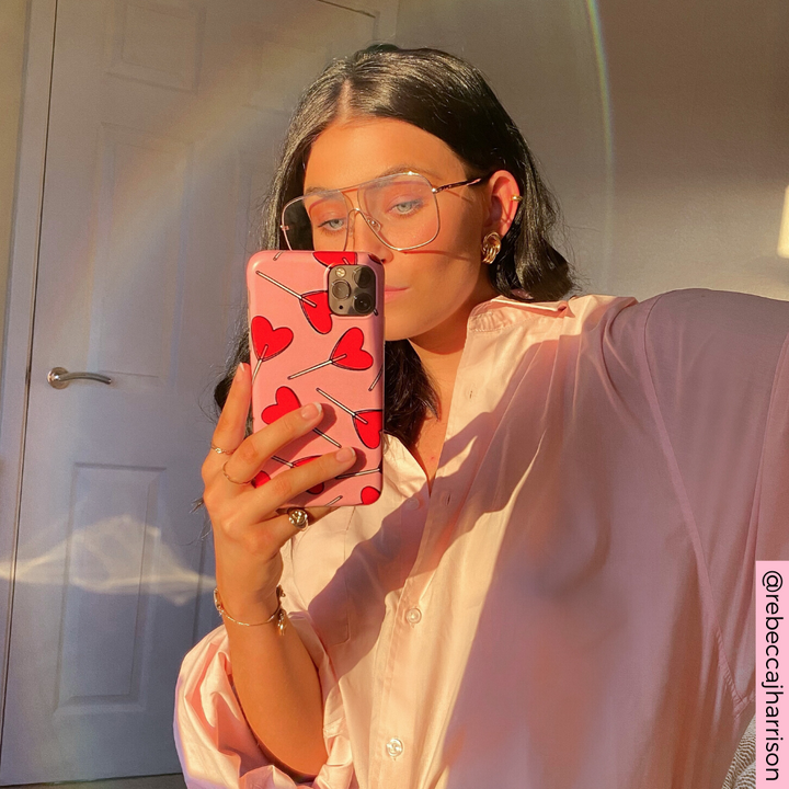 A woman with long black hair takes a mirror selfie with a phone that has a Coconut Lane Candy Hearts Phone Case. She wears a loose, light pink shirt and large, clear-rimmed glasses. Sunlight creates a warm glow and a ring of light reflections in the background.