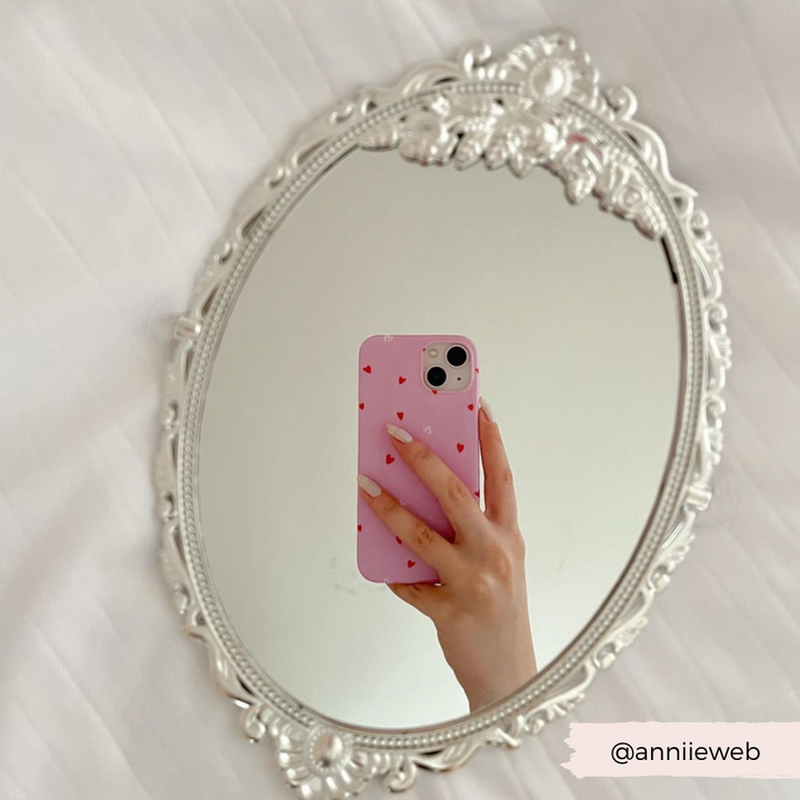 A hand holding Coconut Lane's Cute Hearts Phone Case for the iPhone 15, adorned with an adorable hearts pattern, is reflected in an ornate oval mirror with a silver frame. The background showcases a soft, white textured surface. The Instagram handle @anniieweb is written in the bottom right corner, making it perfect for Valentine's Day inspiration.