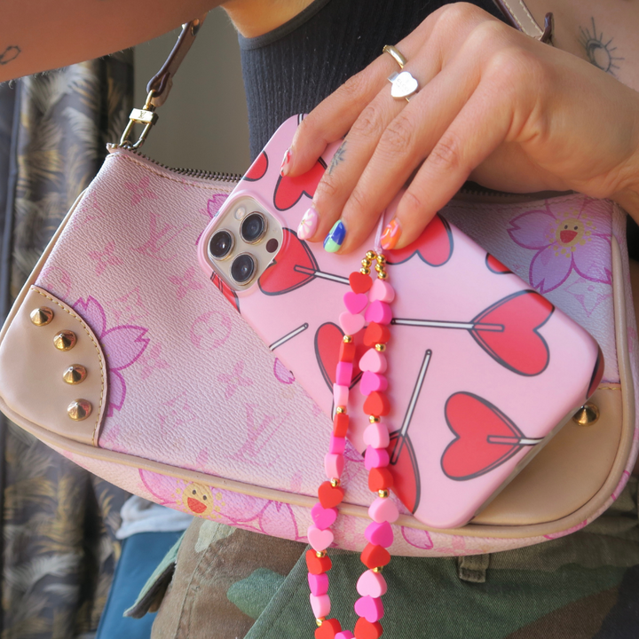 A person holding a smartphone with a pink case adorned with heart designs and the Beaded Phone Strap - Hearts by Coconut Lane. The phone is decorated with a heart-shaped charm. The individual, wearing a ring and showcasing multicolored nails, also carries a pink handbag featuring flower and heart decorations, standing against a patterned background.