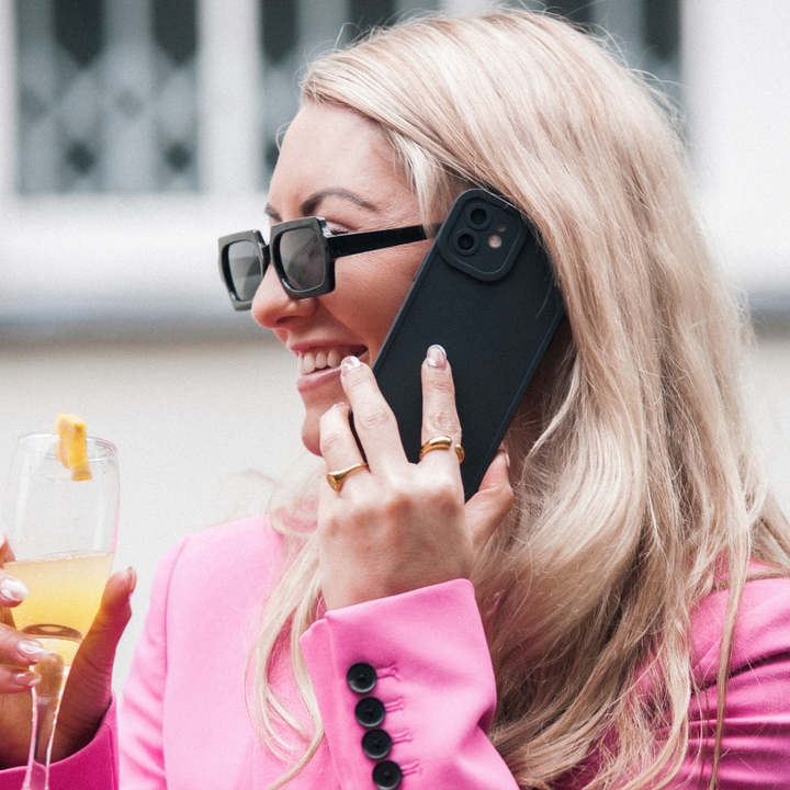 A person with long blonde hair, wearing black sunglasses and a bright pink blazer, is holding a smartphone with a NAKD Phone Case - Midnight Black by Coconut Lane to their ear with one hand while holding a glass of an orange drink with a lemon garnish in the other. They are smiling and appear to be engaged in conversation.
