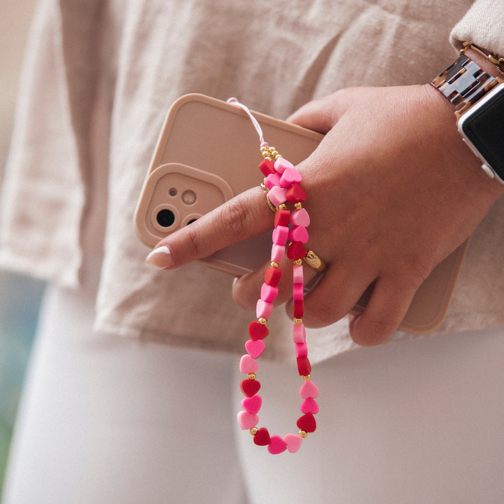 A person holding a smartphone embellished with a beige case and a Coconut Lane Beaded Phone Strap - Hearts, which features small pink, red, and white heart-shaped beads. They are dressed in a neutral-colored top, white pants, and wearing a smartwatch. The phone is comfortably held in their left hand.