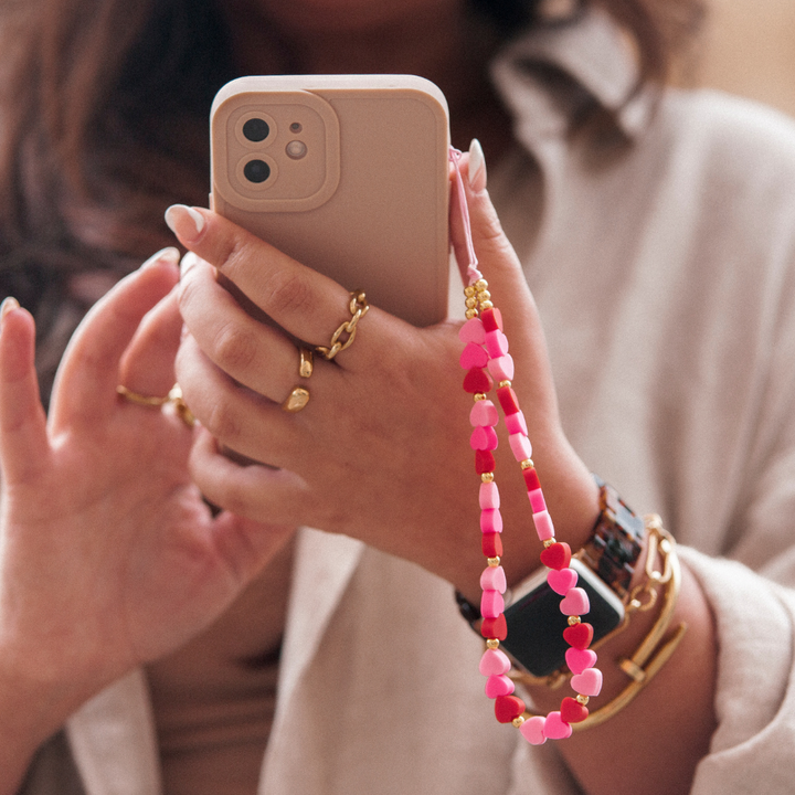 A person with long hair is holding a smartphone in a beige case adorned with Coconut Lane's Beaded Phone Strap - Hearts. They are wearing several gold and black wrist accessories, including a watch and bracelets. The background is blurred, highlighting their hands and the smartphone.