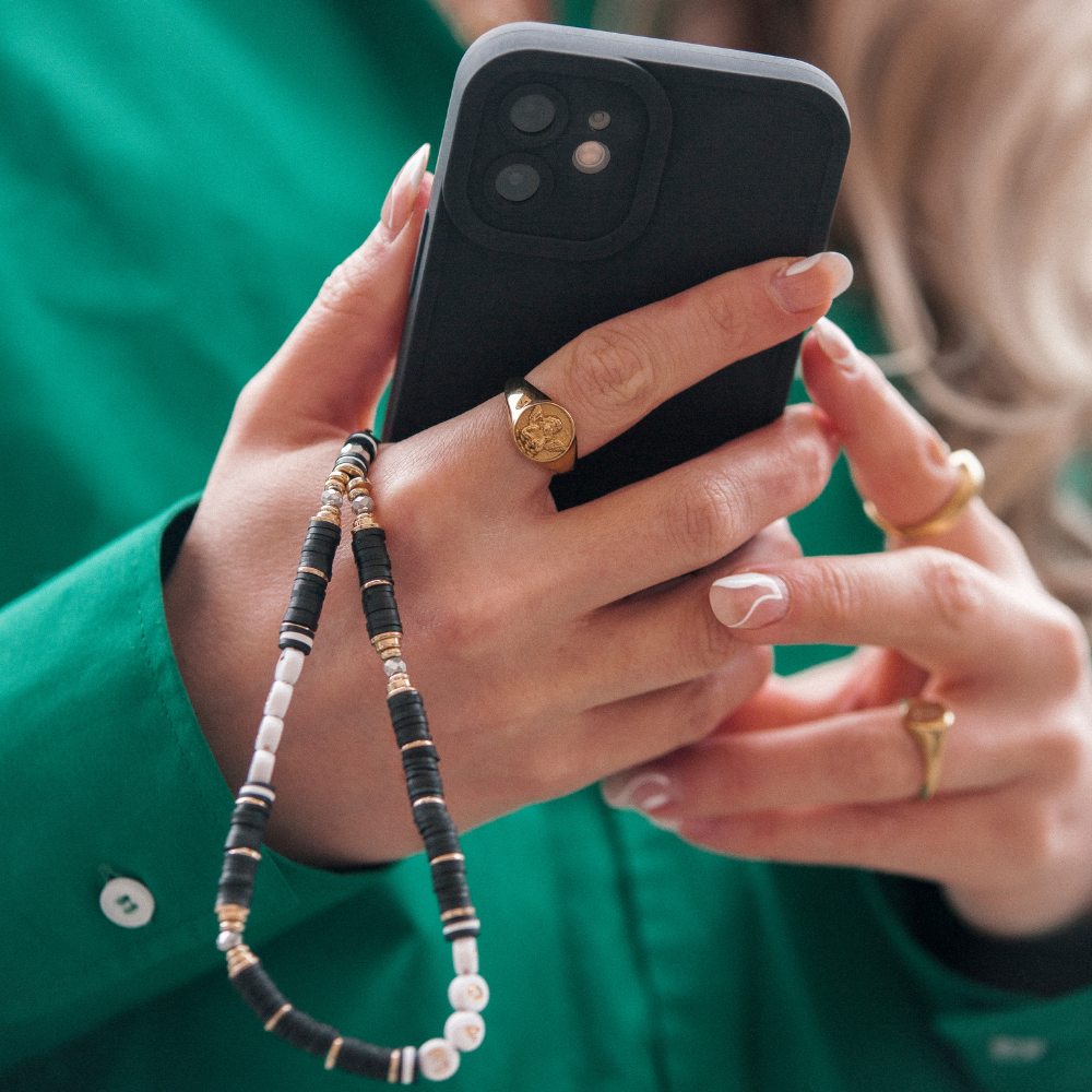 A person wearing a green shirt holds a black smartphone equipped with the NAKD Phone Case - Midnight Black from Coconut Lane, featuring full camera protection and a beaded strap. Their well-manicured nails complement their several gold rings, including one with a detailed symbol. The soft, flexible TPU phone case and strap are adorned with various small beads.