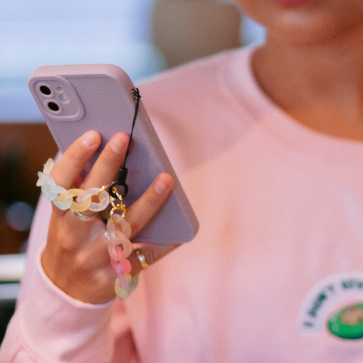 A person wearing a pink sweatshirt from the NAKD spring collection holds their lilac NAKD Phone Case by Coconut Lane, adorned with a colorful chain strap. The phone's full camera protection and finger grip are visible, while the individual's face is partially out-of-frame, concentrating on their pastel favorite.