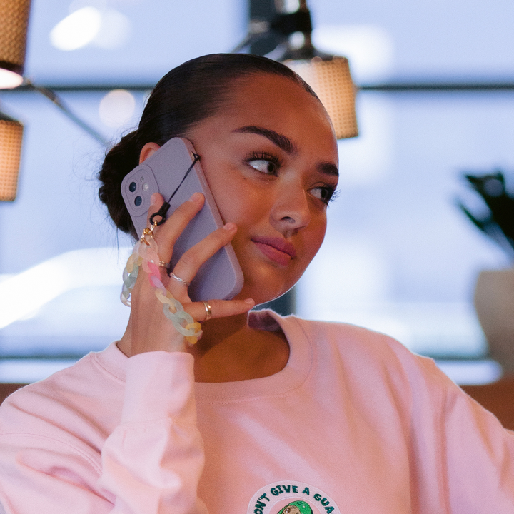 A person with a bun hairstyle is holding a phone in a NAKD Phone Case - Lilac by Coconut Lane to their ear, seemingly in conversation. They are wearing a light pink sweatshirt from the NAKD spring collection and standing indoors, with blurred background elements including lights and plants.
