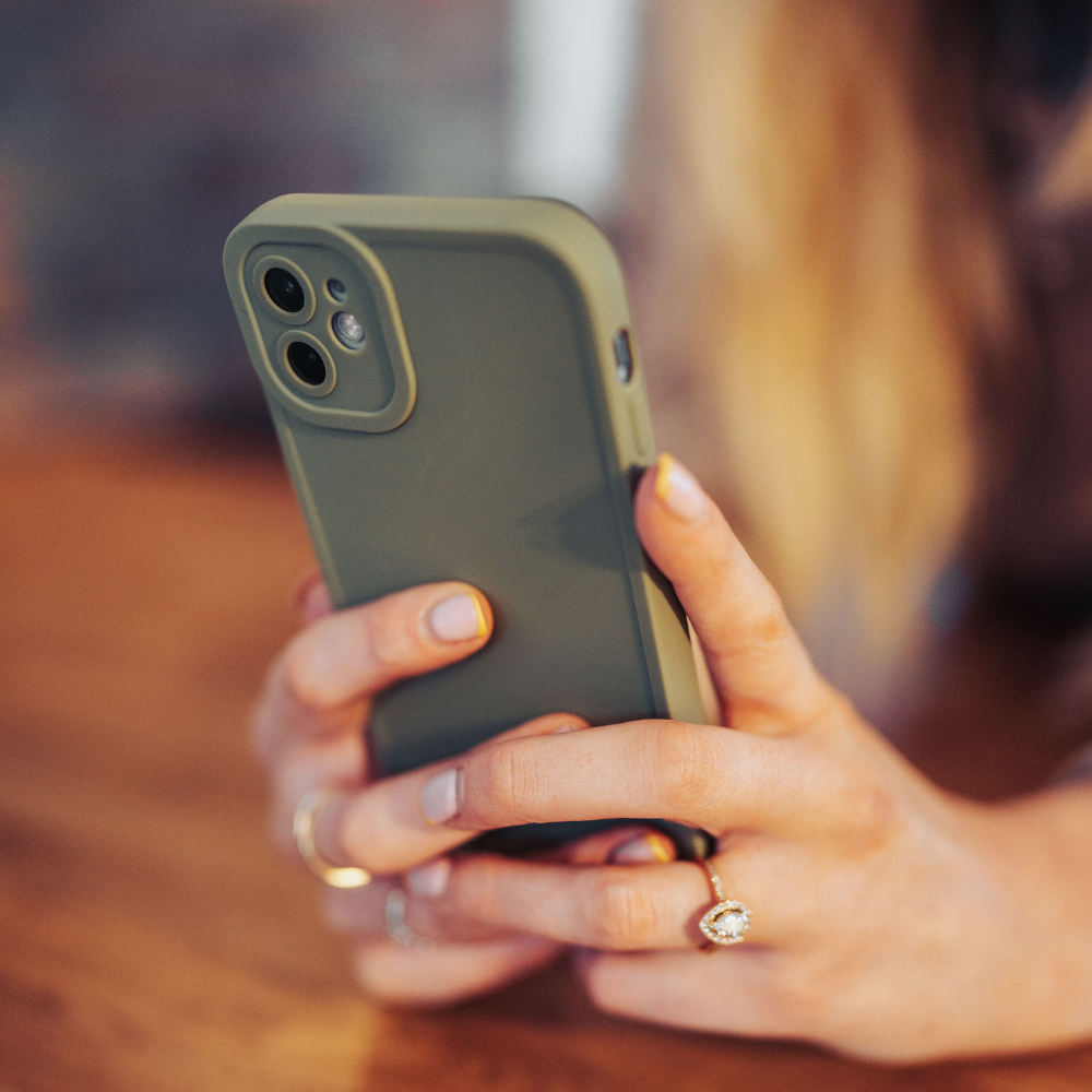 Close-up of a person's hands holding a Coconut Lane NAKD Phone Case - Khaki with full camera protection. The person has long blond hair and is wearing a ring with a heart-shaped stone on one hand. The background is blurred, focusing on the phone and the hands.