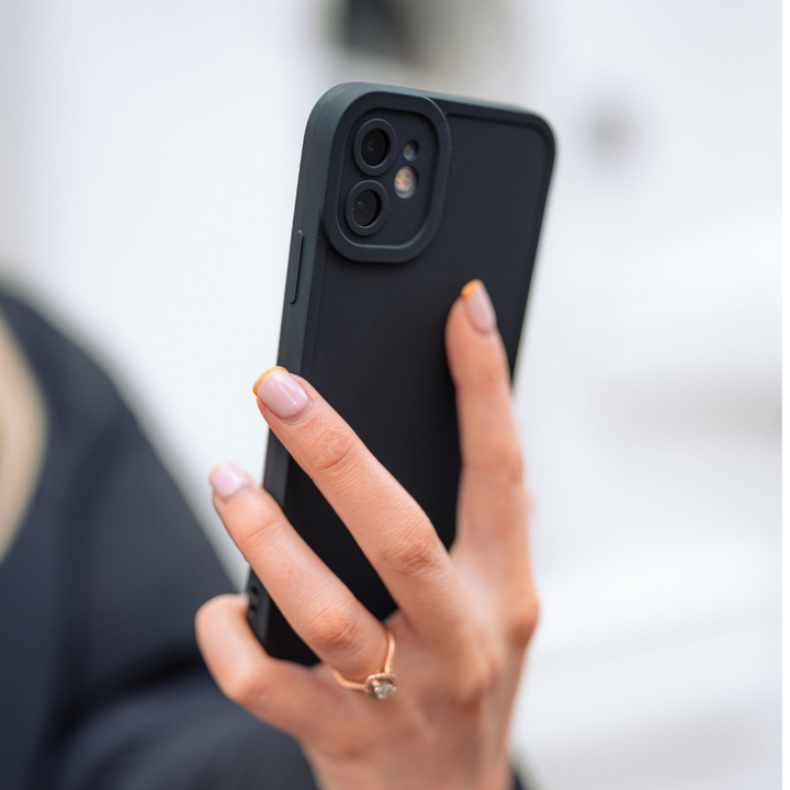 A close-up image of a person holding a black smartphone encased in the Coconut Lane NAKD Phone Case - Midnight Black, which offers full camera protection. The person has light pink nails adorned with yellow tips and wears a ring featuring a small diamond on their ring finger. The background is blurred and white.