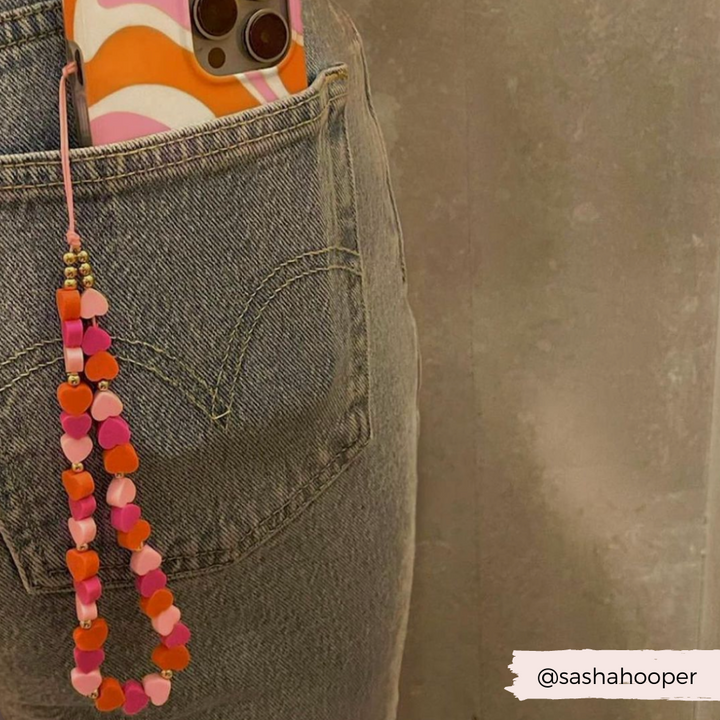 A person is wearing jeans with a phone half tucked into the back pocket. The phone has a colorful case featuring a wavy orange, pink, and white pattern. Attached to the phone is Coconut Lane's Beaded Phone Strap - Hearts, adorned with charming pink and orange heart-shaped beads. Image credit goes to @sashahooper.
