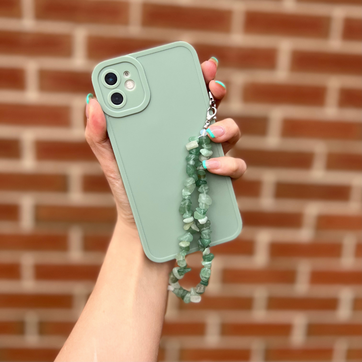 A hand with painted nails holds a green phone with a matching green case. Attached to the phone case is Coconut Lane's Crystal Phone Strap - Green, featuring vibrant beads reminiscent of Green Aventurine. The background is a red brick wall.