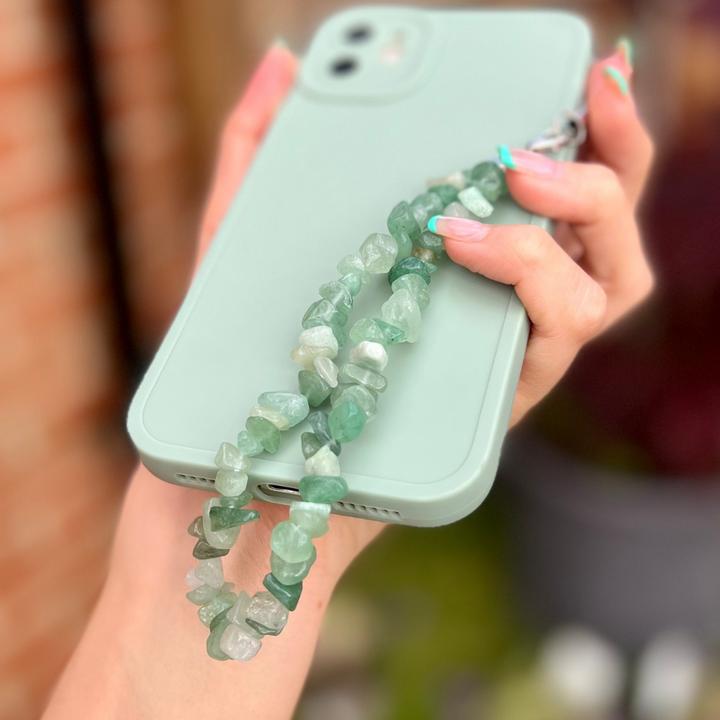 A hand with light-colored nails holds a mint green iPhone encased in a matching mint green cover. Attached to the phone case is the Crystal Phone Strap - Green from Coconut Lane, made of translucent, greenish stones, likely Green Aventurine. The blurred background showcases a mix of greenery and a brick wall.