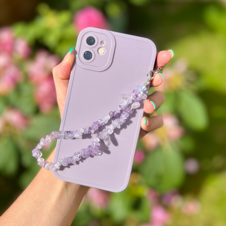 A hand holds a lavender-colored smartphone with the Crystal Phone Strap - Lilac from Coconut Lane attached. The background features blurred greenery and pink flowers, suggesting an outdoor setting. The hand has painted nails in a mix of blue and green colors, highlighting the decorative accessory.