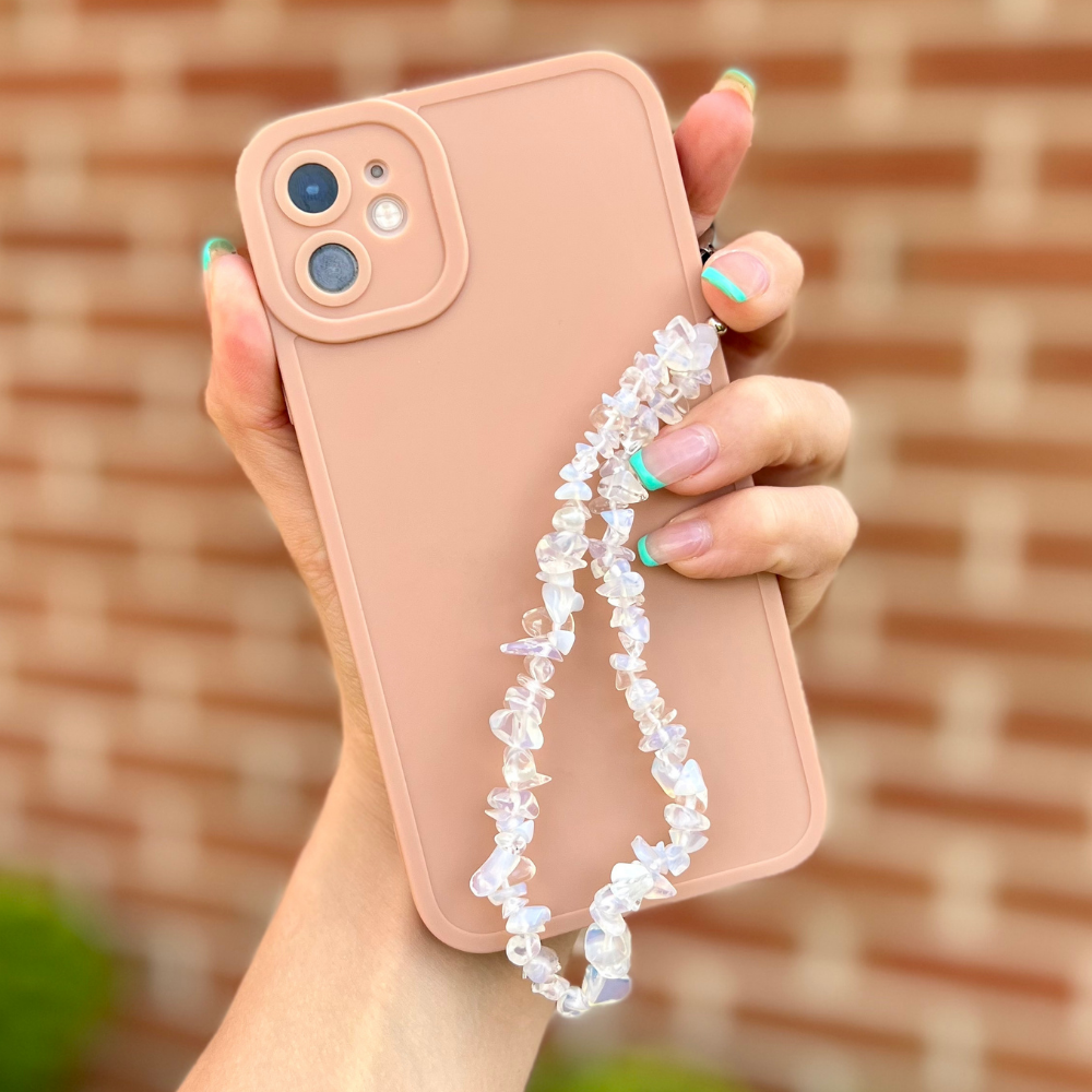 A hand with a French manicure, featuring green tips, holds a smartphone with a light pink NAKD Phone Case - Peach from Coconut Lane. The phone has a beaded phone charm made of translucent stones. The background is a blurred red brick wall.