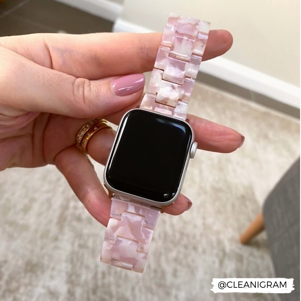A hand with pink-painted nails holds a smartwatch featuring the Luxe Rose Apple Watch Strap by Coconut Lane, characterized by its adjustable resin material and pink marbled design. The band has a glossy finish and a link-style pattern. Gold rings adorn the fingers. The background includes a carpeted floor and part of a chair. "@CLEANIGRAM" is in the corner.