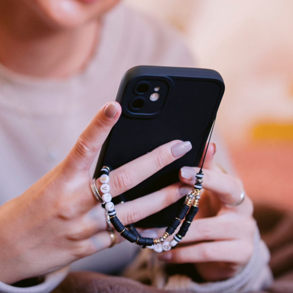 Close-up of a person holding a black smartphone with both hands, featuring the NAKD Phone Case - Midnight Black by Coconut Lane. The phone is adorned with a beaded charm displaying various colorful and patterned beads. The individual has manicured nails and is wearing silver rings on both hands. The background appears blurred.