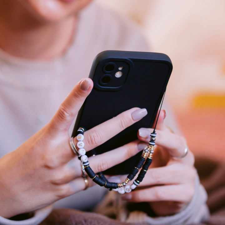 Close-up of a person holding a black smartphone with both hands, featuring the NAKD Phone Case - Midnight Black by Coconut Lane. The phone is adorned with a beaded charm displaying various colorful and patterned beads. The individual has manicured nails and is wearing silver rings on both hands. The background appears blurred.