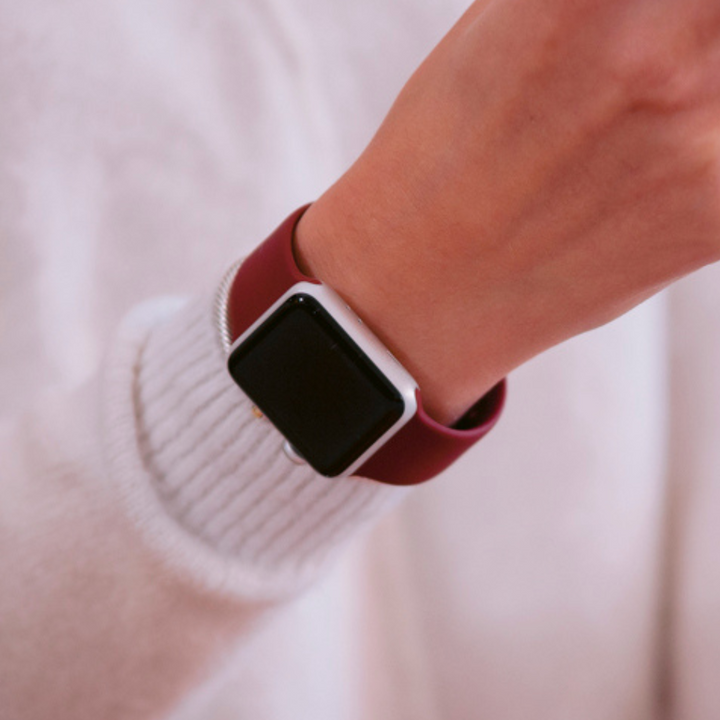 A person wearing a smartwatch with a NAKD Apple Watch Strap - Red Wine by Coconut Lane on their wrist. The background is blurred, and the individual is dressed in a white sweater. The smartwatch screen is turned off.