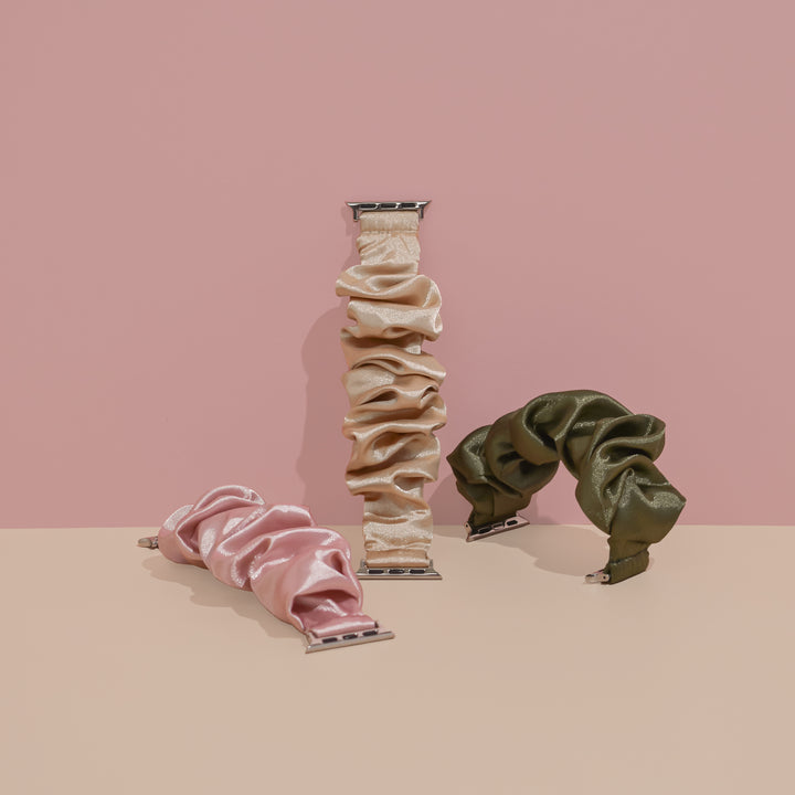 Three silky satin Scrunchie Apple Watch Straps from Coconut Lane are displayed against a two-toned pink and beige backdrop. The straps, in elegant colors of dusty pink, beige, and olive green, showcase a stylish Apple Watch accessory. The beige strap stands upright in the center with the dusty pink and olive green straps on either side.
