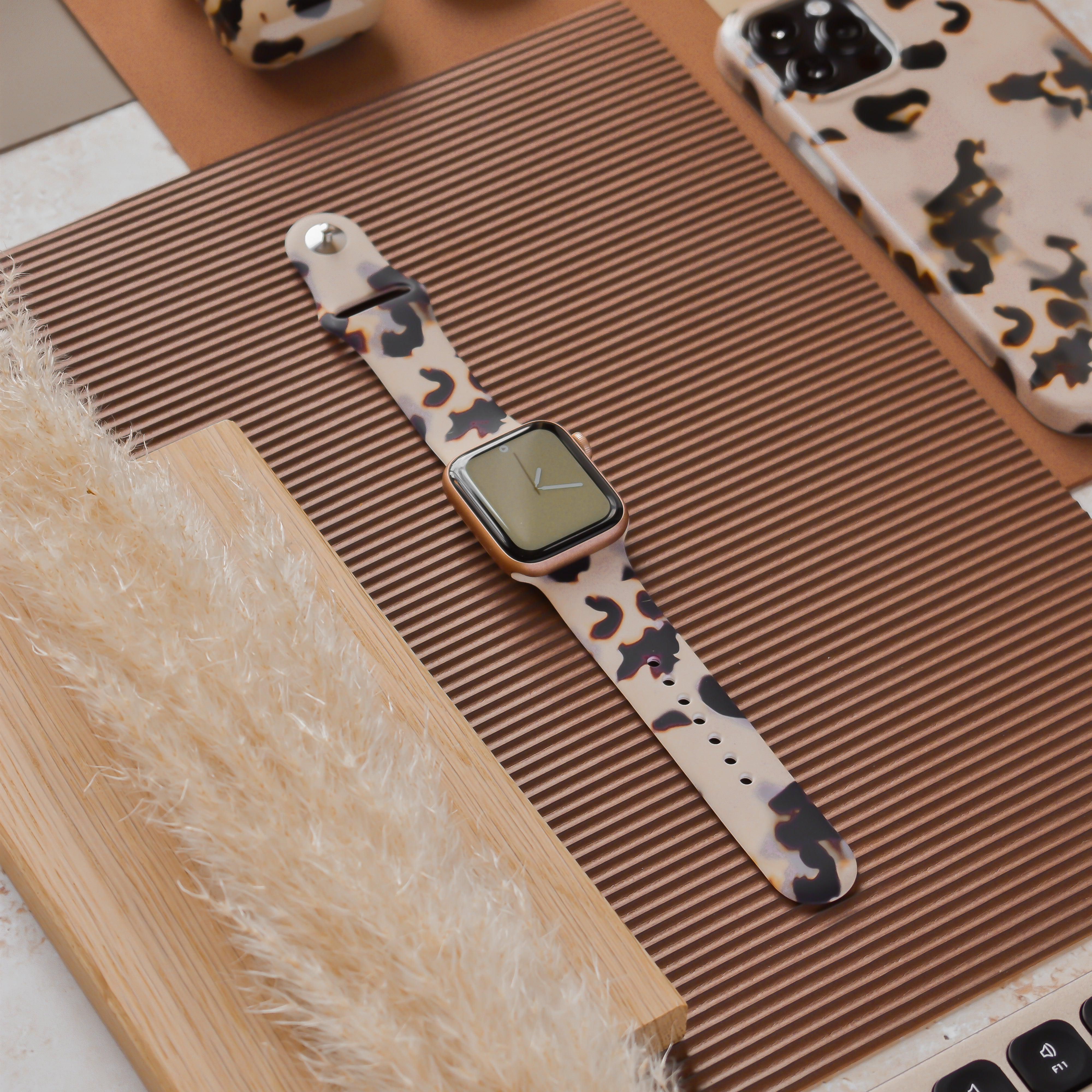 A smartwatch with a Coconut Lane Milky Ivory Tort Apple Watch Strap is placed on a brown ribbed mat. Surrounding it are matching beige and black camouflage accessories, including a phone case and a pair of headphones. A fluffy beige decoration is also partially visible.