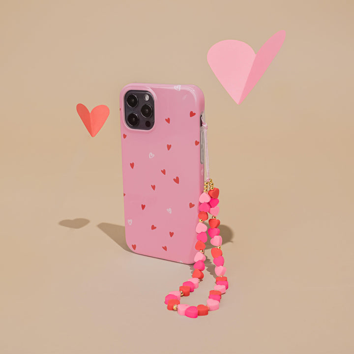 A pink phone case adorned with small red and white hearts stands upright against a beige background. Attached to the case is Coconut Lane's Beaded Phone Strap - Hearts in pink and red. Two heart-shaped paper cutouts in red and pink float nearby.