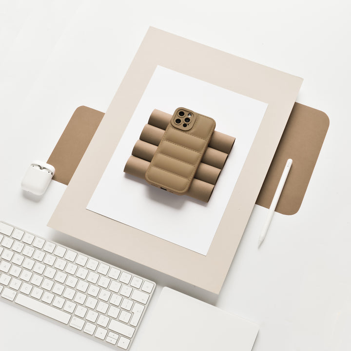 A minimalist desk setup features a modern iPhone with a Quilted Phone Case - Brown by Coconut Lane, resting on a stack of foam rollers. Nearby, a white Apple Magic Keyboard, a pencil, and a closed AirPods case hint at a clean and organized workspace. All items are arranged on a white surface.