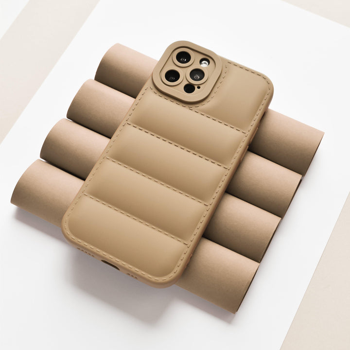 The Quilted Phone Case - Brown by Coconut Lane is displayed lying face down on a set of brown cylindrical objects, showcasing its full camera protection. Made from soft, flexible TPU, the background features a neutral palette with white, beige, and light gray tones.