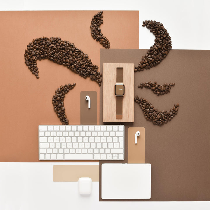 A flat lay image features an Apple Watch with a Coconut Lane Magnetic Chain Apple Watch Strap in Coffee color on a wooden stand, accompanied by a white wireless keyboard, white wireless earbuds with a charging case placed on tan and brown sheets, and coffee beans arranged artfully to resemble eyebrows, eyes, and smiling lips.