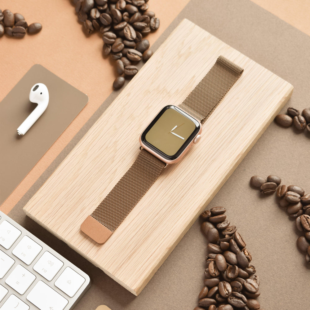 A sleek smartwatch adorned with the Coconut Lane Magnetic Chain Apple Watch Strap in Coffee, featuring a stylish brown mesh band and stainless steel magnetic clasp, rests on a wooden surface surrounded by scattered coffee beans. Nearby, there is a wireless earbud, part of a keyboard, and a mouse pad, all set against a beige and brown background.