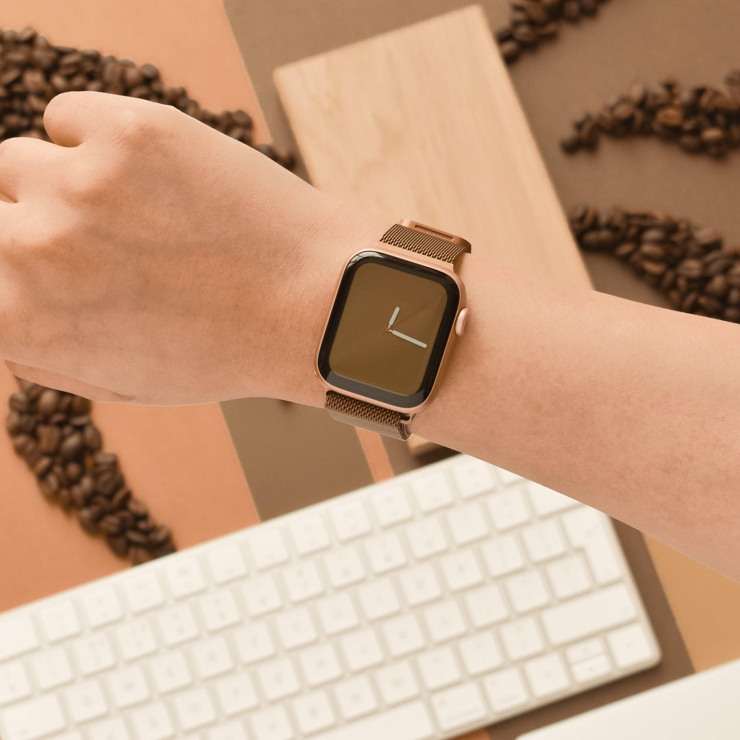 Close-up of a person's wrist wearing an Apple Watch with a minimalist clock face and a Coconut Lane Magnetic Chain Apple Watch Strap - Coffee. The background features a white computer keyboard and scattered coffee beans on a brown surface.