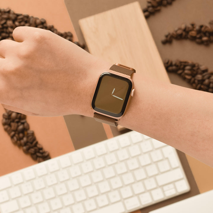 Close-up of a person's wrist wearing an Apple Watch with a minimalist clock face and a Coconut Lane Magnetic Chain Apple Watch Strap - Coffee. The background features a white computer keyboard and scattered coffee beans on a brown surface.