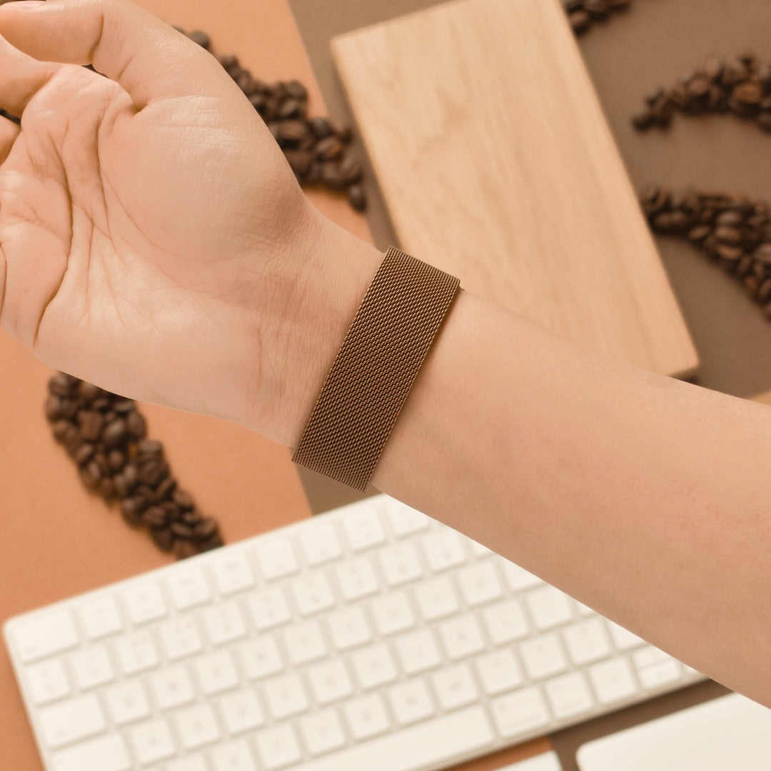 A hand wearing the Coconut Lane Magnetic Chain Apple Watch Strap in Coffee is shown over a beige and brown background. Coffee beans, a white computer keyboard, and a wooden block are in the background.