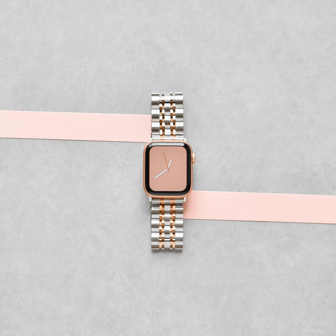 A Stainless Steel Apple Watch Strap by Coconut Lane, featuring a silver and rose gold metal link band, is placed on a flat surface. The watch face is rose gold, displaying the time around 10:00. A light pink ribbon runs horizontally beneath the watch, adding a touch of elegance to the adjustable strap for a perfect fit.