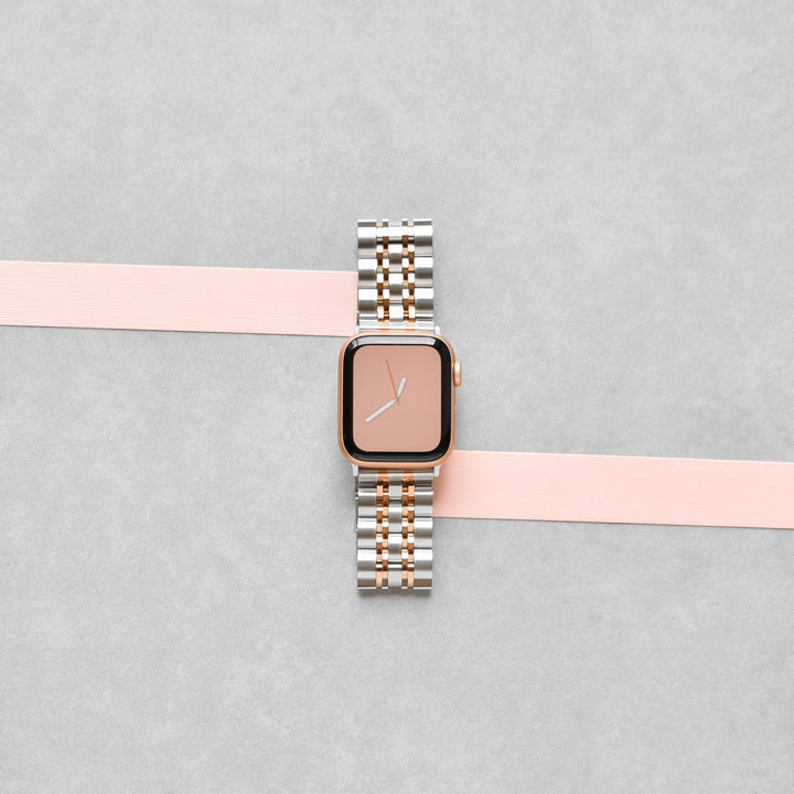 A Stainless Steel Apple Watch Strap by Coconut Lane, featuring a silver and rose gold metal link band, is placed on a flat surface. The watch face is rose gold, displaying the time around 10:00. A light pink ribbon runs horizontally beneath the watch, adding a touch of elegance to the adjustable strap for a perfect fit.