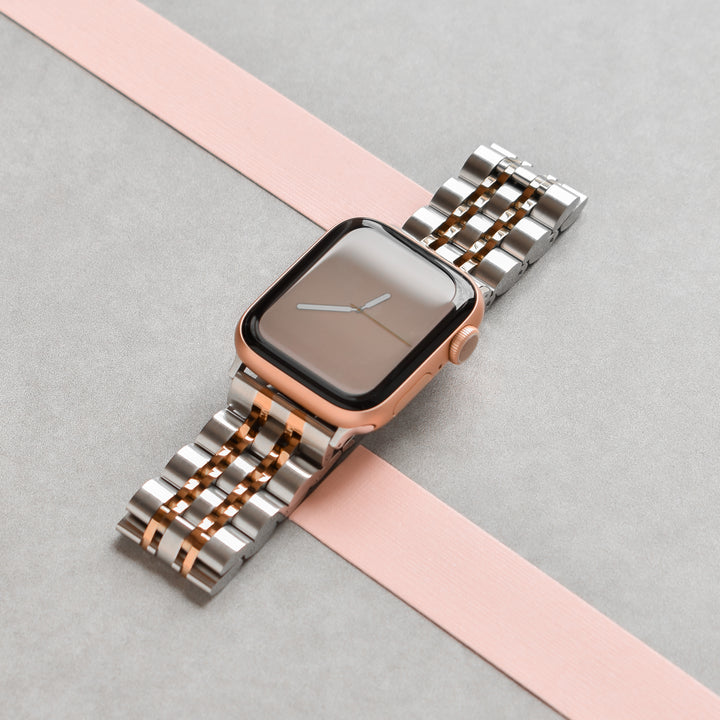 A Coconut Lane Stainless Steel Apple Watch Strap - Silver & Rose Gold is displayed on a two-tone background of grey and pink, attached to a smartwatch with a rectangular face. The watch shows the hands of an analog timepiece but appears to be turned off or in sleep mode.