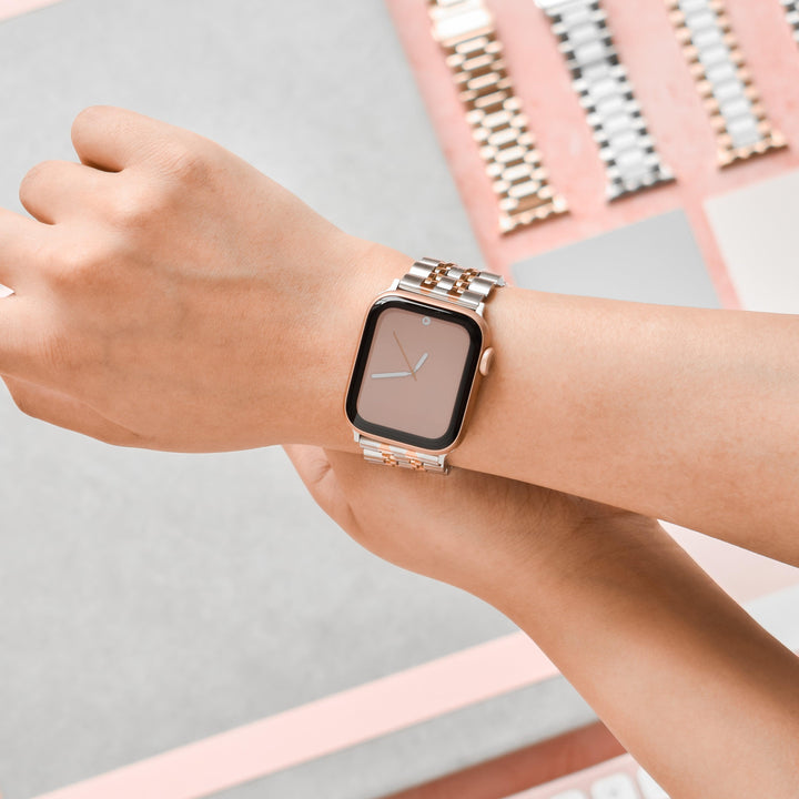 A person is wearing a Coconut Lane Stainless Steel Apple Watch Strap in Silver & Rose Gold on their left wrist. The background features various metal watch bands in different styles and colors, neatly arranged on a light pink surface. The person appears to be adjusting the watch with a metal band link remover tool.