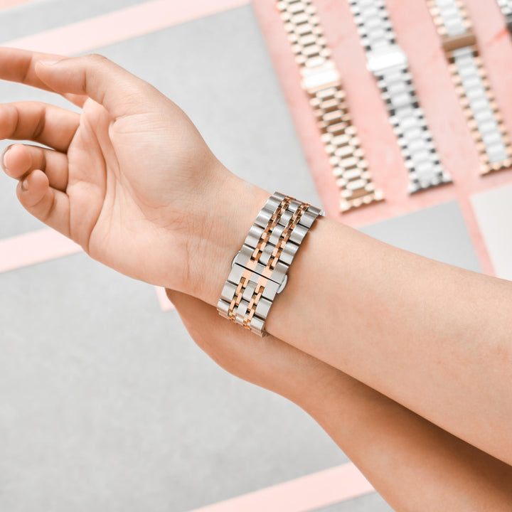 Close-up of a person trying on a Stainless Steel Apple Watch Strap in Silver & Rose Gold by Coconut Lane. The person's wrist and hand are shown, with additional Stainless Steel Apple Watch Straps displayed in the background on a table. The straps feature varied finishes, predominantly in silver and rose gold tones.