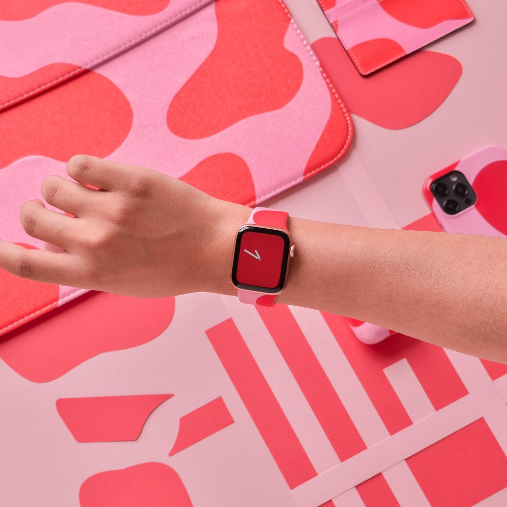 A wrist adorned with Coconut Lane's Abstract Pink & Red Apple Watch Strap. The background is filled with various red and pink patterned items, including a laptop sleeve, phone case, and fabric swatches, all harmonizing in matching colors and designs. The overall aesthetic is modern and vibrant.