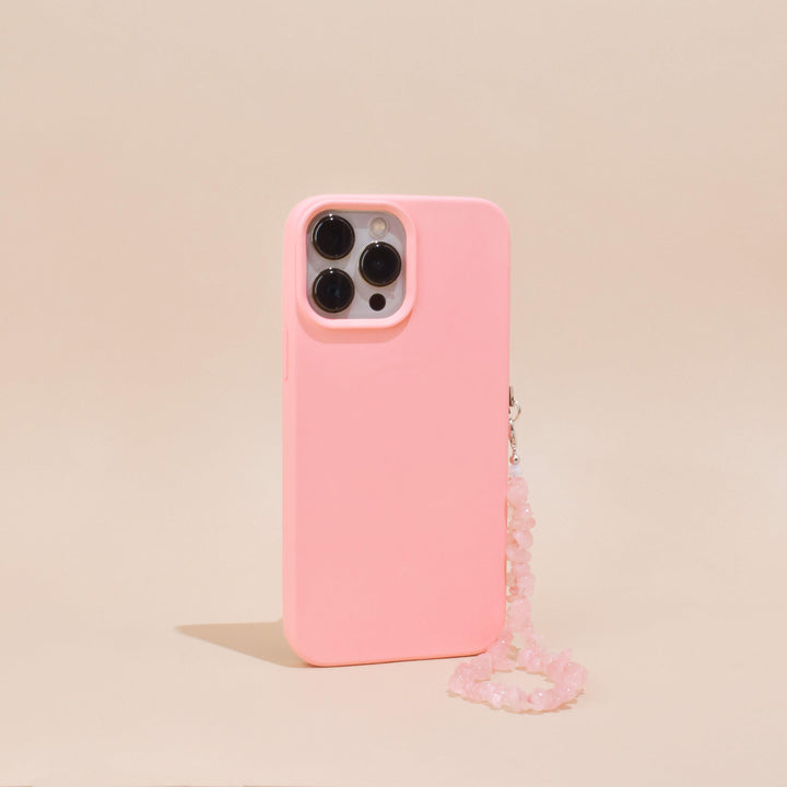 A pink smartphone case featuring Coconut Lane's Crystal Phone Strap - Rose Quartz is displayed on a beige background. The case features precise cutouts for three camera lenses and a flash. The pink beaded strap, adorned with genuine rose quartz crystals, is attached to the side of the case, providing an elegant decorative touch.