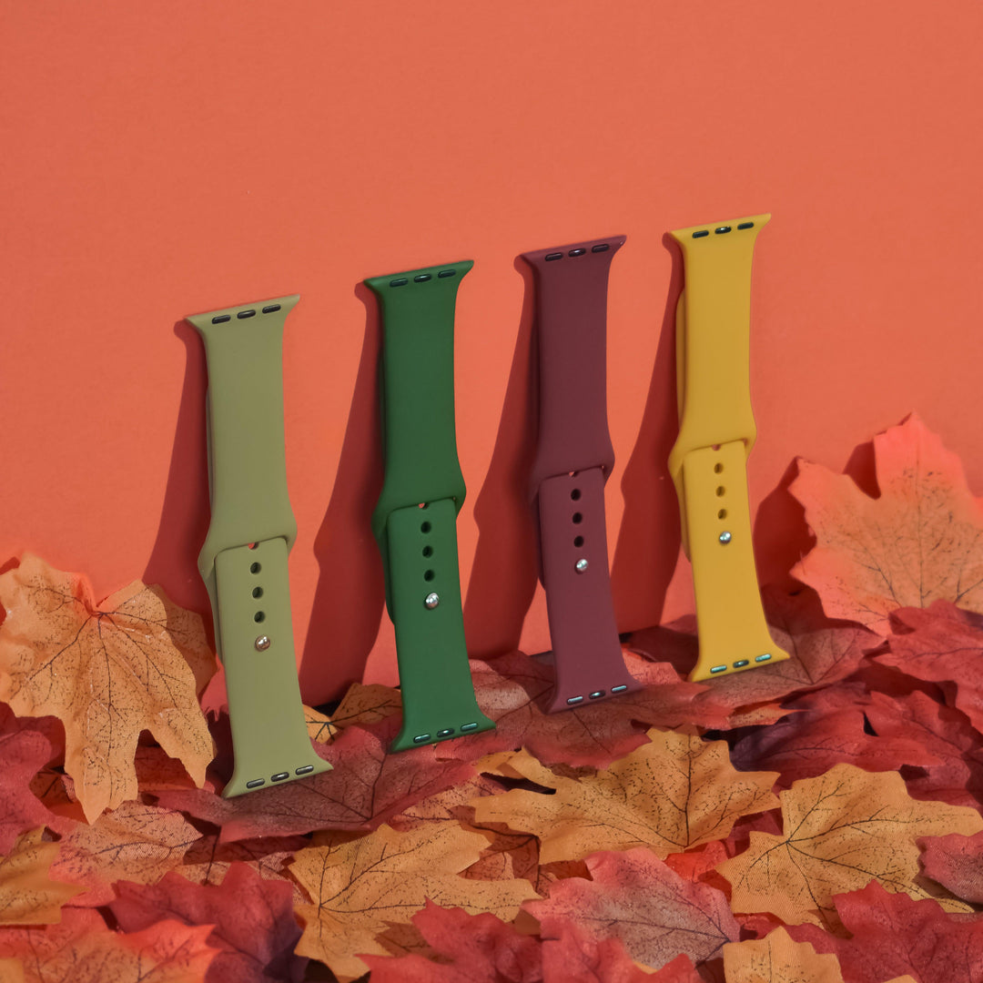 A display from the Coconut Lane NAKD Collection showcases four smartwatch bands in autumn colors—light green, dark green, burgundy, and mustard yellow—standing upright against an orange background and surrounded by scattered autumn leaves. It features the NAKD Apple Watch Strap in Olive Green, made of durable silicone.