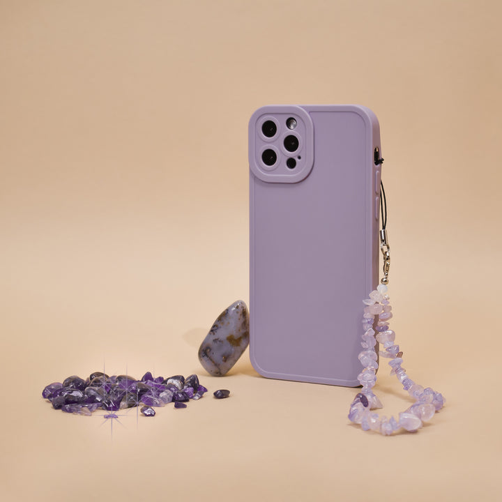 A phone case in a vibrant lilac shade with Coconut Lane's Crystal Phone Strap adorned with small beads stands upright against a beige background. Scattered around the stylish accessory are amethyst crystals and one larger, smooth stone with earthy tones.