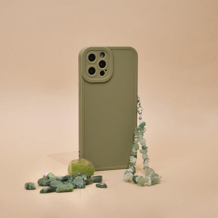 A Crystal Phone Strap - Green by Coconut Lane is displayed on a beige background, showcasing its beaded charms attached to a green phone case. Small green stones and a polished Green Aventurine gemstone are scattered beside it, creating a minimalist and earthy aesthetic.
