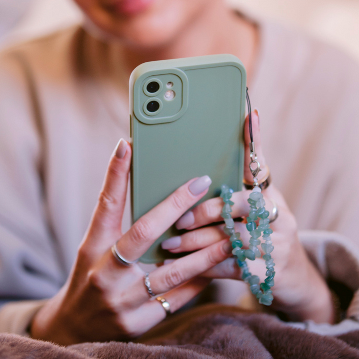 A person with manicured nails is holding a green smartphone adorned with the Crystal Phone Strap - Green from Coconut Lane. They are wearing a light-colored shirt and several rings on their fingers. The background is blurred, emphasizing the hands and the phone.