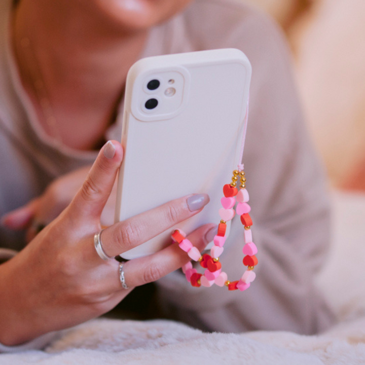 A person holding a white smartphone adorned with Coconut Lane's Beaded Phone Strap - Hearts, featuring pink and red beads. The person is dressed in a beige top, and a silver ring can be seen on their finger. The background is blurred to keep the focus on the smartphone and the charming phone strap.