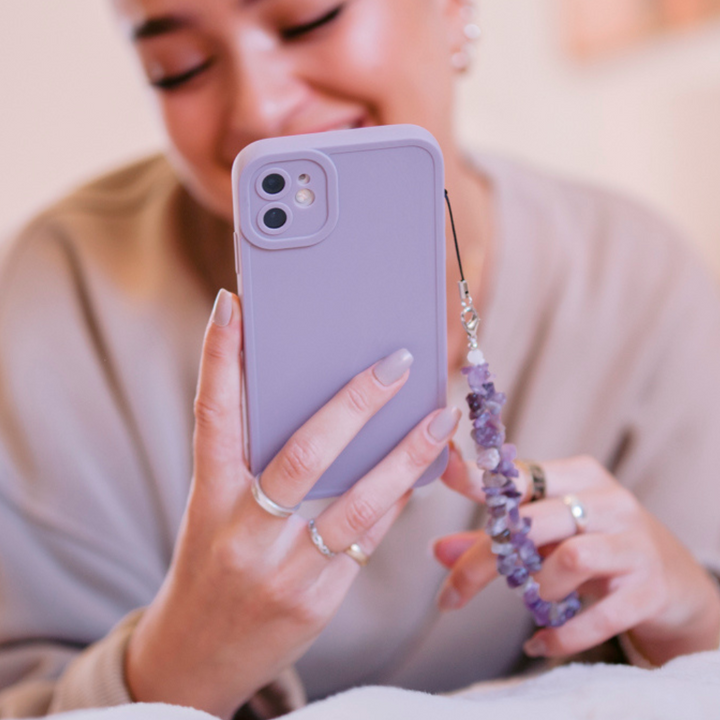 A person smiles while holding a smartphone with a lavender-colored case and decorative accessory. Their fingers, adorned with rings, also flaunt Coconut Lane's Crystal Phone Strap - Lilac on a beaded phone strap attached to the case. The background is softly blurred.
