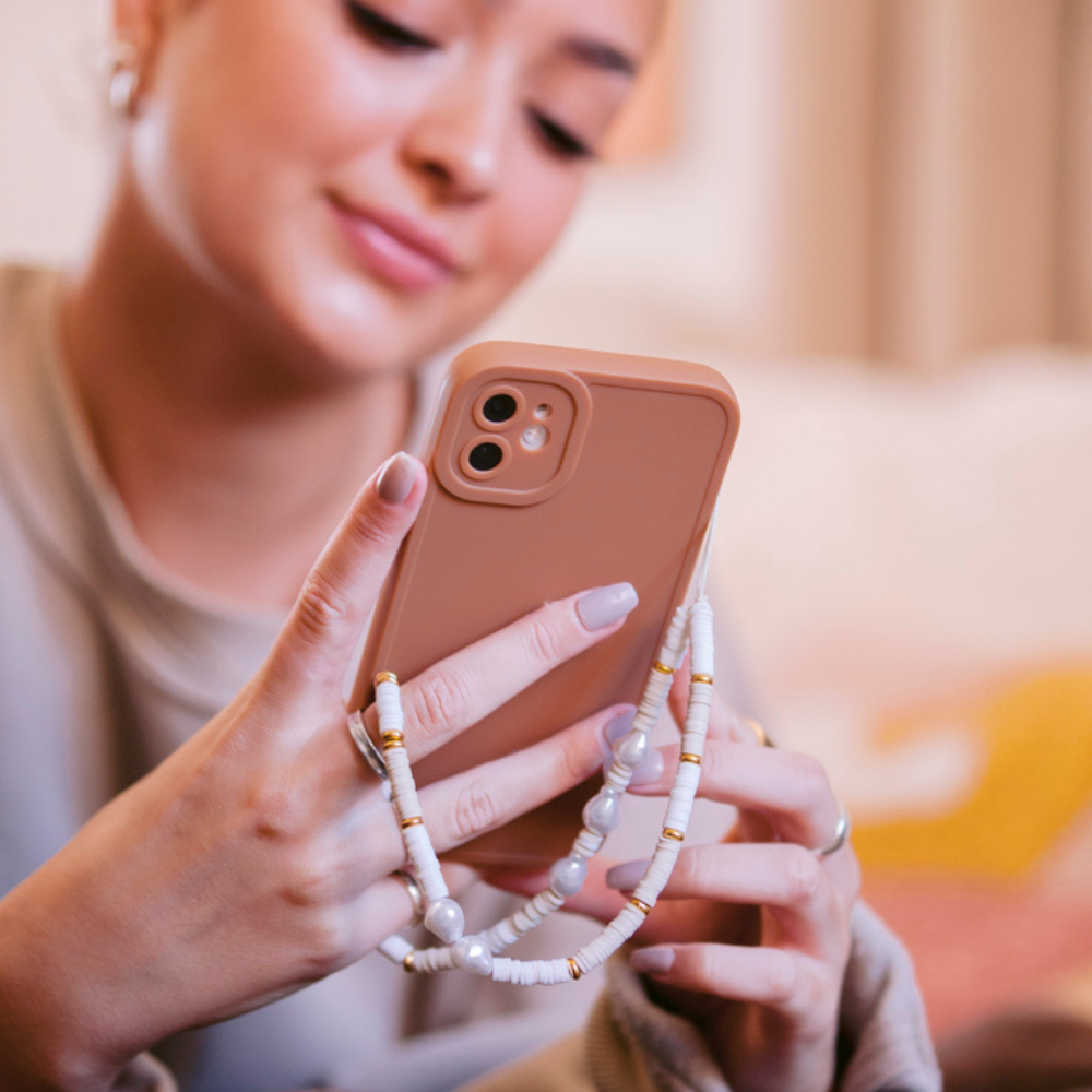 A person with light-colored nails holds a pink smartphone adorned with a beaded strap. The background is softly blurred, and the individual appears to be seated and engaged with the phone, smiling slightly, while a NAKD Phone Case - Chocolate Brown from Coconut Lane rests beside them.