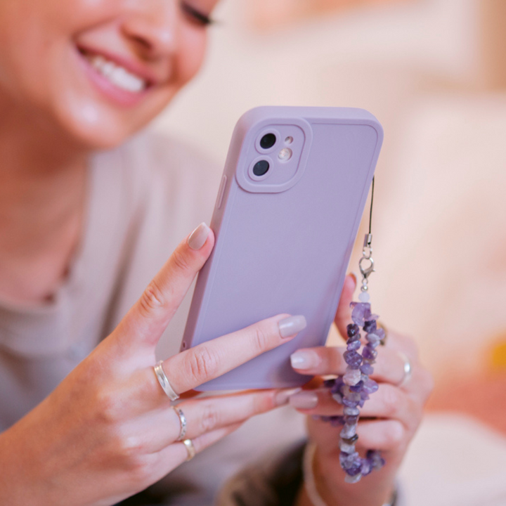 A person with a smile is holding a smartphone with a lavender-colored case and the Crystal Phone Strap - Lilac from Coconut Lane, which features decorative accessories and amethyst crystals. The focus is on the phone, while the person's face appears slightly blurred in the background. The person has manicured nails and is wearing several rings.